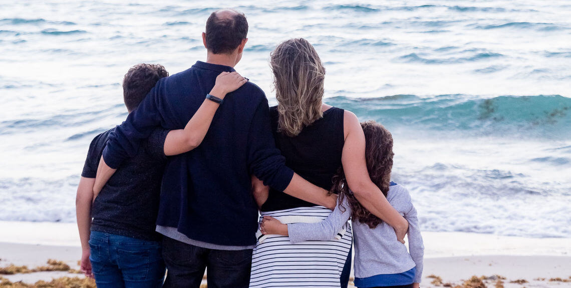 Familie steht zusammen am Strand und schaut aufs Meer